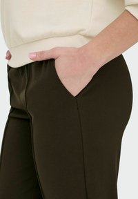 Mörkgröna byxor med sidofickor, slät textur och en åtsittande design, kombinerat med en krämfärgad, ribbad sweatshirt.