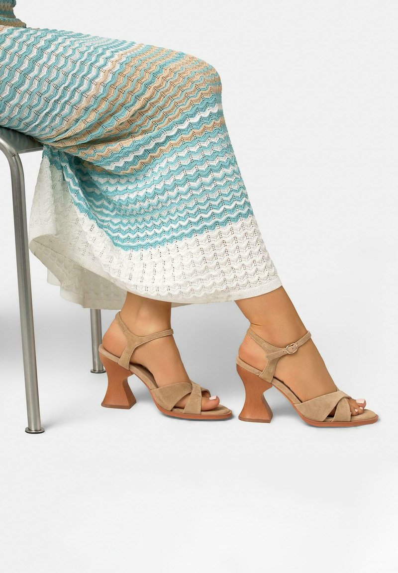 Femme portant des sandales à talons en daim beige avec talons blocs courbés et une jupe multicolore en crochet, assise sur une chaise en métal.