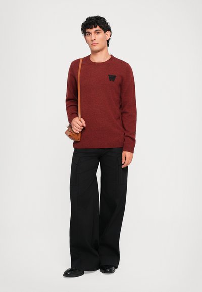 Maglione a maglia bordeaux con scollo rotondo e logo nero, abbinato a pantaloni neri a gamba larga. Il modello porta una borsa a tracolla marrone.