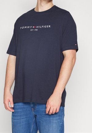 Mann trägt ein marineblaues Tommy Hilfiger T-Shirt mit Logo und dem Schriftzug "EST. 1985", kombiniert mit blauen Denim-Jeans.