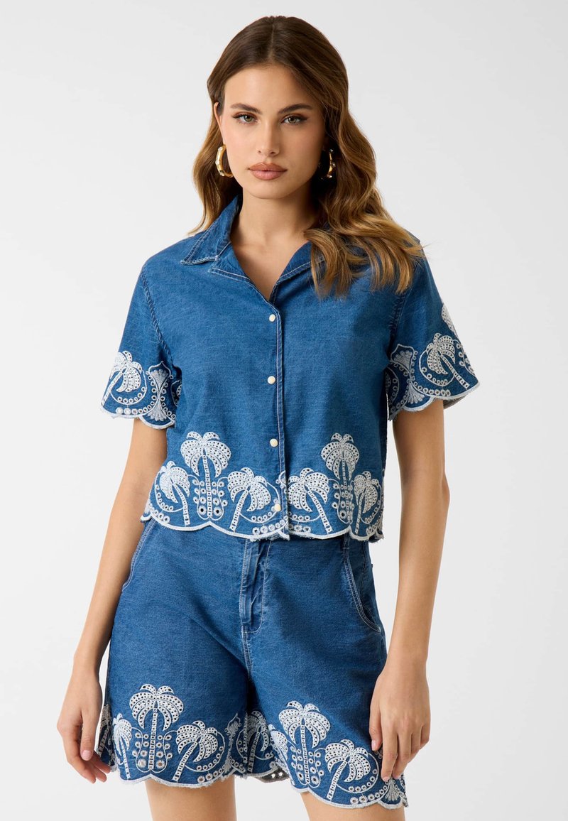 Femme portant un ensemble chemise à manches courtes et short en denim bleu avec des broderies de palmiers blancs sur les bords, debout devant un fond uni.