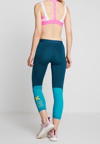 Kvinna som bär en rosa sport-bh och turkosa leggings med färgblock och gula och vita bokstäver, stående barfota mot en enkel grå bakgrund.