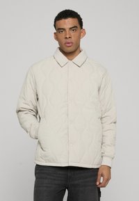 Kockás bézs kabát, gallérral, visszafogott hullámos mintákkal, patent gombokkal és bordázott mandzsettákkal. Sötét farmernadrág fölött viselve.