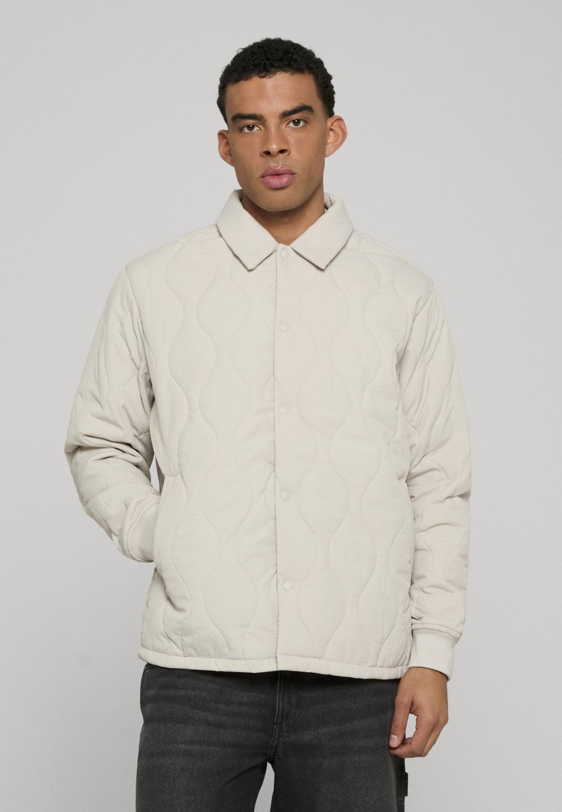 Kockás bézs kabát, gallérral, visszafogott hullámos mintákkal, patent gombokkal és bordázott mandzsettákkal. Sötét farmernadrág fölött viselve.