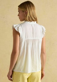 Weiße, leichte Bluse mit Rüschen-Ärmeln, gerafftem Rückendetail und Rundhalsausschnitt. Tragen mit hellen Hosen.