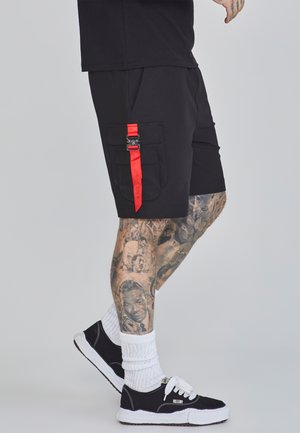 Pantalones cortos de carga negros con dos bolsillos laterales, que presentan un detalle de correa roja y hebilla. Combinados con zapatillas en blanco y negro y calcetines blancos.