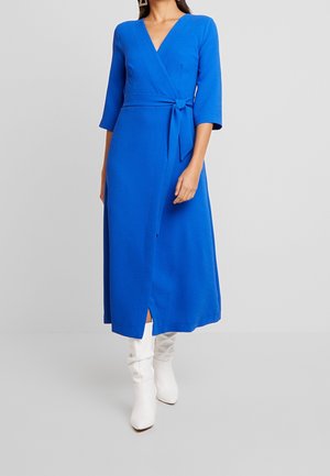 Femme portant une longue robe portefeuille bleu roi à manches trois-quarts et des bottes blanches mi-mollet, debout devant un fond uni.