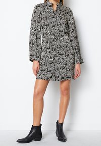 Schwarz-weiß gemustertes Kleid mit langen Ärmeln, Kragen, Taillengürtel und geradem Schnitt. Getragen mit kniehohen schwarzen Stiefeletten.