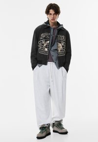 Musta lyhyt takki graafisella painatuksella, yllä harmaa ruudullinen paita. Valkoiset väljät housut ja harmaat saappaat, joissa vihreät nauhat.