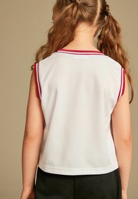 Ærmeløs hvid jersey-top med røde detaljer, der har en tekstureret mesh-stof og en løs pasform, parret med mørkegrønne shorts.