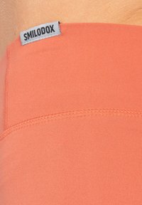 Zbliżenie na koralowy materiał z przyszytym szwem i szarym metkiem oznaczonym "SMILODOX" na skórze osoby.