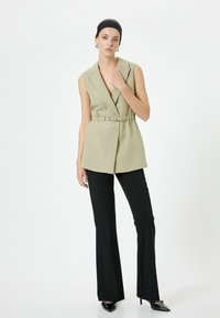 Khaki ärmellose Blazer mit einem Gürtel, kombiniert mit schwarzen Schlaghosen und spitzen Absatzschuhen, vor einem weißen Hintergrund.