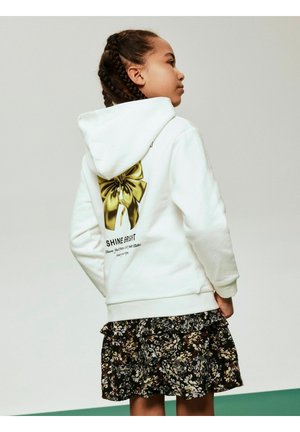 Witte hoodie met een goudkleurige strikafbeelding en de tekst "SHINE BRIGHT" op de achterkant, gecombineerd met een donker bloemenruche rok.