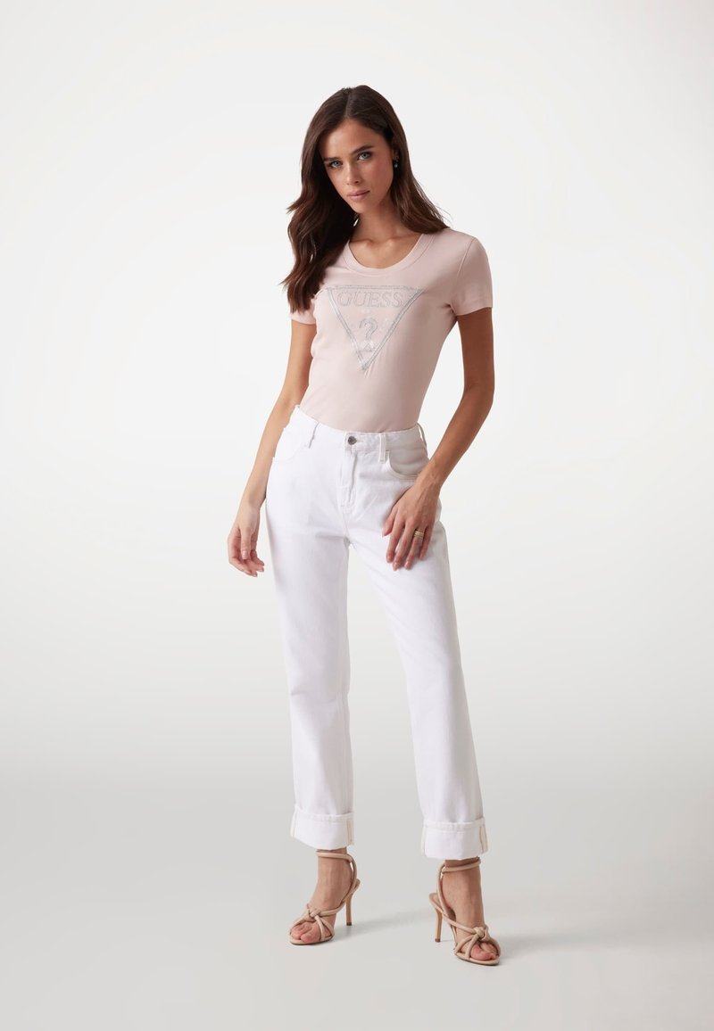 Camiseta ajustada de color rosa claro con un logo triangular plateado, combinada con jeans de mezclilla blancos con dobladillo y sandalias de tacón beiges. Textura suave en general.