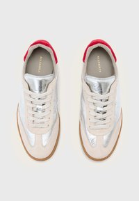 Beige színű velúr testű, ezüst metál panelekkel és piros részletekkel a saroknál lábbeli. Lapos fűzőkkel és beige talpbetéttel rendelkezik.