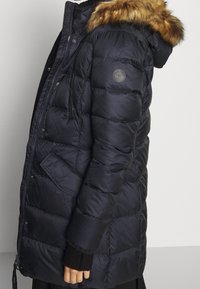 Marineblauwe pufferjack met een nepbont capuchon, gevoerd patroon, ritssluiting en zijzakken. Bevat een logo op de mouw.