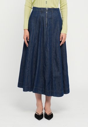 Femme portant une jupe midi en jean bleu foncé taille haute avec une fermeture éclair à l'avant, associée à un haut vert clair à manches longues côtelé et des ballerines noires à bouts pointus.