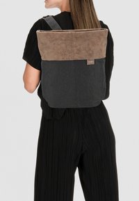 Mochila con una parte superior de cuero marrón y un cuerpo de tela negra. Presenta correas ajustables y un cierre de cremallera. Se observa un branding mínimo.