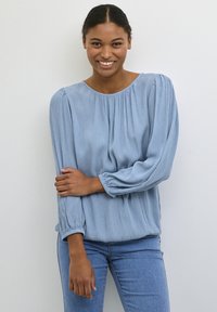 Helles blaues Langarmbluse mit gerafftem Kragen, strukturiertem Stoff und subtilen erhabenen Mustern. Kombination mit blauen Jeans.
