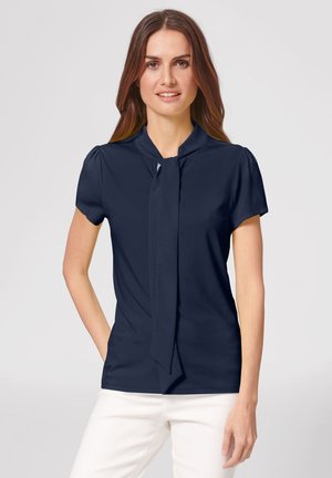 Frau trägt eine marineblaue kurzärmelige Bluse mit einer vorderen Schleife am Hals, kombiniert mit weißen Hosen, vor einem hellgrauen Hintergrund stehend.