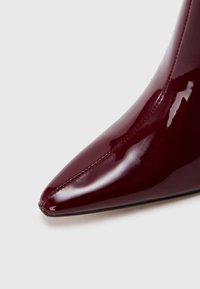 Botte à talon haut bordeaux brillant à bout pointu avec couture visible sur une surface en cuir lisse, sur fond blanc.