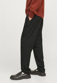 Pantalon noir à coupe ample, avec des poches latérales et une texture lisse. Porté avec des mocassins noirs, complété par un pull rouge.