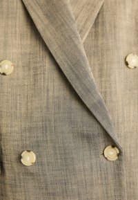Beige kaksirivinen bleiseri, joka on valmistettu teksturoidusta kankaasta, jossa on hienovarainen herringbone-kuvio. Varustettu pyöreillä vaaleilla nappeilla ja leveillä kauluksilla.