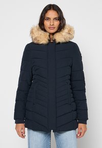 Giacca imbottita blu navy con cappuccio rifinito con pelliccia sintetica, design trapuntato e due tasche laterali. Tessuto liscio con silhouette aderente.