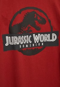 Röd bomullströja med en svart siluett av en dinosaurie inom en cirkulär logotyp. Texten lyder "JURASSIC WORLD DOMINION" i vitt.