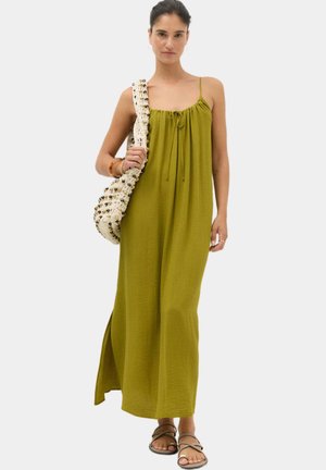 Kvinde iført en olivengrøn ærmeløs maxikjole, bærende en stor strikket beige taske og iført brune sandaletter med remme.