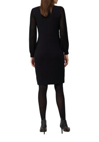 Femme portant une robe noire à mi-longueur avec des manches longues transparentes, des collants noirs et des bottines à talons noirs, de dos par rapport à l'appareil photo.