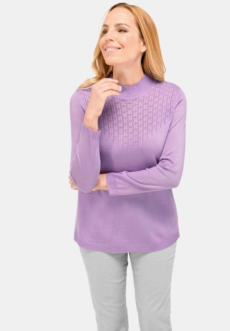 Lavendelfarbenes Strickpullover mit hohem Hals und strukturiertem Muster auf der oberen Brust; weicher Stoff und lange Ärmel. Kombiniert mit hellgrauen Hosen.