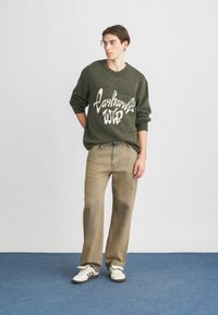 Pulover verde tricotat cu text grafic alb, asortat cu pantaloni drepți maro deschis și adidași albi cu dungi negre.