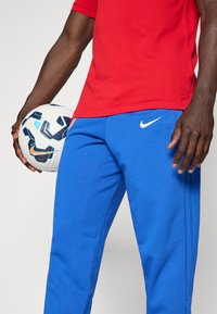 Red short-sleeve shirt paired with blue Nike sweatpants, featuring white logo accents and a textured white soccer ball with blue details held in hand.