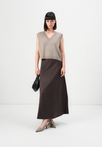 Donna con capelli scuri e lisci, che indossa un top senza maniche beige lavorato a maglia, una lunga gonna marrone scuro e tacchi beige, mentre tiene una piccola borsa nera, in piedi su uno sfondo bianco.