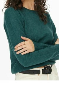 Maglione teal con scollatura rotonda, polsini a costine e una consistenza morbida. Indossato con una cintura in pelle nera sopra pantaloni chiari.