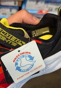 Scarpa nera Skechers con accenti gialli, dotata di etichetta di chiusura adattiva. Fodera in mesh multicolore con suola imbottita e design aerodinamico.