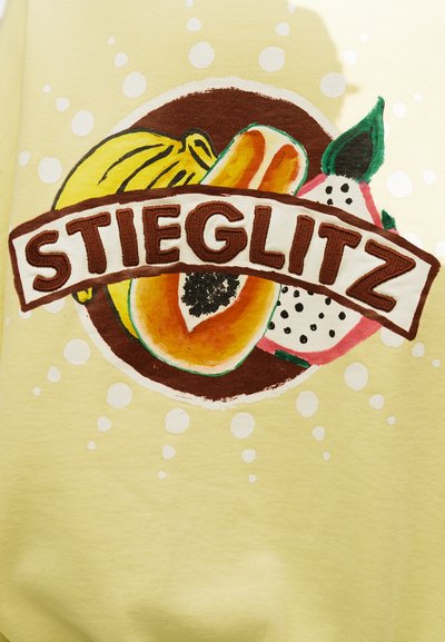 Tissu jaune avec des illustrations de fruits colorés derrière le texte "STIEGLITZ" en lettres brunes en gras, entouré de pois blancs sur un fond clair.