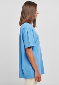 Femme aux cheveux bruns lisses, de profil, portant un T-shirt ample bleu clair et un pantalon blanc, sur fond gris uni.