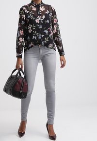 Blouse noire transparente avec motif floral, jean skinny gris, et sac à main avec accents bordeaux. Note : des escarpins à bout pointu complètent la tenue.