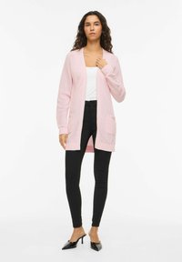 Világos rózsaszín kötött kardigán V-nyakkivágással, nyitott elejű, hosszú ujjas, két első zsebbel, fekete szűk farmernadrággal és fekete magassarkú cipővel párosítva.