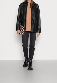 Persona che indossa una giacca di pelle nera, un maglione di cotone beige, jeans neri, stivali neri e tiene in mano una piccola borsa a tracolla a strisce zebrate.