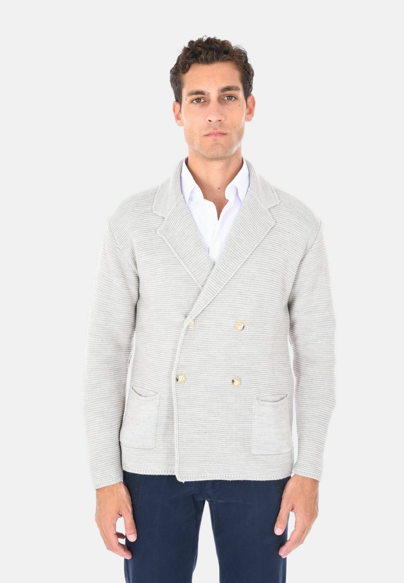 Giacca a maglia doppio petto di colore grigio chiaro, impreziosita da bottoni dorati, colletto a risvolto e due tasche frontali, abbinata a una camicia bianca sotto.
