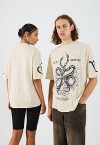 Krémové oversized tričko s černým grafickým potiskem znázorňujícím hadí motiv. Krátké rukávy s dekoračními prvky na rukávech.