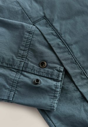 Dark teal fabric with a smooth texture features a folded cuff and two black snap buttons, highlighting tailored design details.