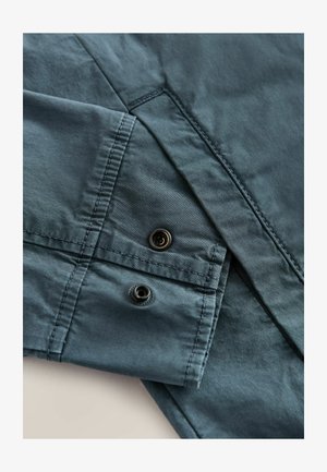 Dark teal fabric with a smooth texture features a folded cuff and two black snap buttons, highlighting tailored design details.