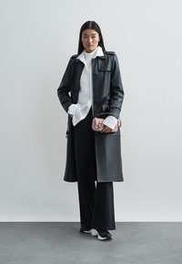 Dugi crni kožni kaput s gumbima, nošen preko bijele bluze i crnih širokih hlača. Mala ružičasta torbica drži se sa strane.