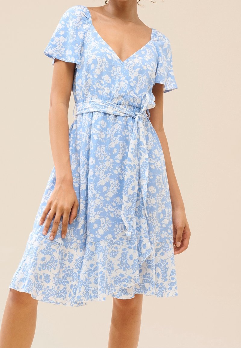 Mujer con un vestido floral azul claro de mangas cortas, escote en V, cinturón atado a la cintura y dobladillo con volantes, sobre un fondo beige.