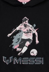 Fußballspieler, der im Actionpose den Ball tritt, mit "MESSI" und einem stilisierten "M"-Logo darunter auf schwarzem Stoff.