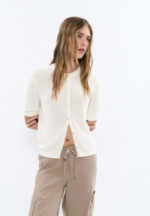 Giovane donna con lunghi capelli castano chiaro indossa un cardigan bianco a manica corta con bottoni e pantaloni beige con coulisse, in piedi con le braccia dietro la schiena.